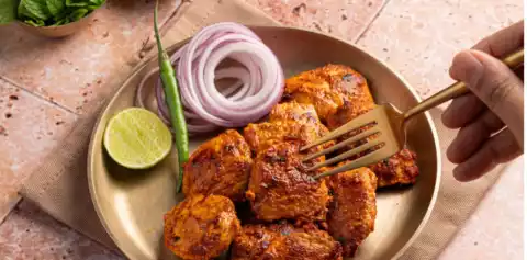 Golden-brown chicken tikka pieces on a brass plate with onion rings, a lime half and a green chilli, a fork lifting one piece